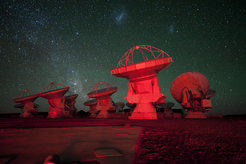 A number of ALMA antennae bathed in red light seen against the Chajnantor night sky. In the background there is the southern Milky Way on the left and the Magellanic Clouds at the top.