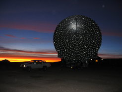 Pathfinder backlit by the sun just below the horizon at dusk: The Atacama Pathfinder Experiment had already begun operation in the Atacama Desert in Summer 2005. With an antenna diameter of 12 metres, it was the largest submillimetre telescope in the Earth’s southern hemisphere. The camera flash is reflected off the adjustment screws of the individual panels, which permit the surface of the dish to be very precisely set.