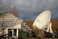 Der Besucherpavillon des Radio-Observatoriums Effelsberg mit dem 100-m-Radioteleskop im Hintergrund. Dort finden auch in diesem Jahr in den Monaten Juli bis Oktober Vorträge für Besuchergruppen statt. Aufgrund der Vorgaben durch das Corona-virus sind aber sowohl die maximale Anzahl der Vorträge pro Tag als auch die Teilnehmerzahl eingeschränkt.
Vom Pavillon aus führt ein kurzer Weg bis zu einem Aussichtspunkt unmittelbar vor dem großen Reflektor. Bitte beachten Sie, dass elektronische Geräte, vor allem Mobiltelefone, in Reichweite des Teleskops komplett ausgeschaltet oder in Flugmodus gebracht werden sollten, da die ausgesandte Strahlung („Elektrosmog“) die Messungen mit dem Radioteleskop empfindlich beeinträchtigt.