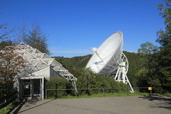Abb. 1: Der Besucherpavillon des Radio-Observatoriums Effelsberg mit dem 100-m-Radioteleskop im Hintergrund. Dort finden in den Monaten April bis Oktober Vorträge für Besuchergruppen statt. Vom Pavillon aus führt ein kurzer Weg bis zu einem Aussichtspunkt unmittelbar vor dem großen Reflektor. Bitte beachten Sie, dass elektronische Geräte, vor allem Mobiltelefone, in Reichweite des Teleskops komplett ausgeschaltet oder in Flugmodus gebracht werden sollten, da die ausgesandte Strahlung („Elektrosmog“) die Messungen mit dem Radioteleskop empfindlich beeinträchtigt.