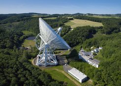 Abb. 1: Das Radio-Observatorium Effelsberg im Luftbild. In der Bildmitte das 100-m-Radioteleskop, recht davon das Observatoriumsgebäude und vorne rechts die neue Werkhalle. Links hinter dem großen Teleskop ist das LOFAR-Antennenfeld zu sehen (Effelsberg-Station des europäischen Niederfrequenz-Radioteleskops LOFAR), eine von sechs Anlagen dieser Art in Deutschland. Der weiße „Fleck“ rechts oberhalb des Teleskops zeigt das Dach des Besucherpavillons. Vom Pavillon aus führt ein kurzer Zickzack-Weg bis zu einem Aussichtspunkt unmittelbar vor dem großen Reflektor.