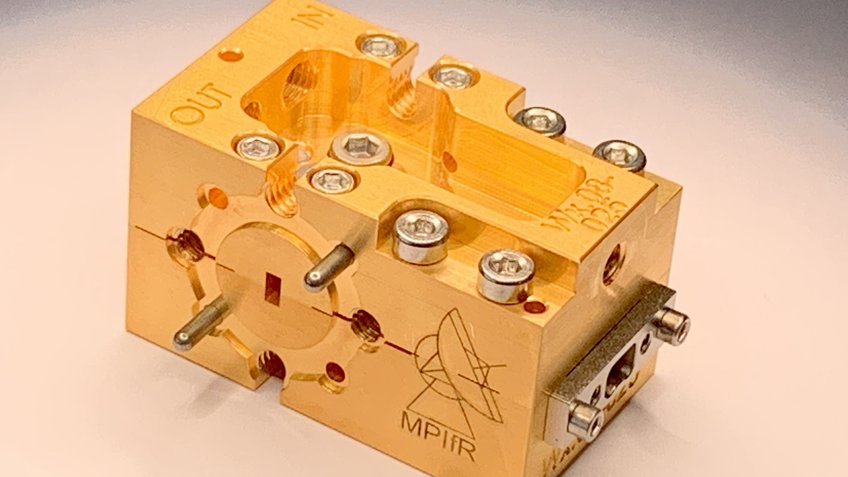 ALMA Band 2 LNA A rectangular gold-coloured device with a port at the front. The MPIfR logo is engraved on the side. A number of screws and openings for screws can be seen all over the device. At the back, there is an engraving saying "out" on the left and an engraving saying "in" on the right.