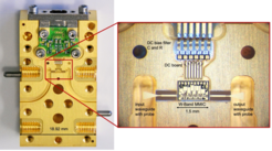 On the left ist a golden device with a great number of holes for screws, On the right, a section in the uper middle of the device is highlighted and zoomed in on. The object is labelled "W-band MMC" and is marked as being 1.5 mm in width. To the left and right there are two waveguides with probe, one labelled input and one output. Above. the W-band MMC, we see a DC board and above that DC bias filters C and R, all labelled accordingly.